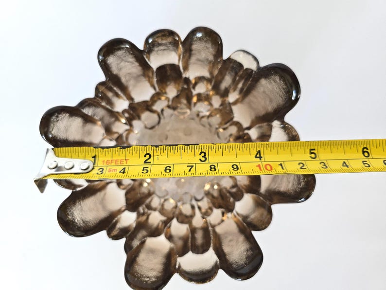 May include: A brown glass vase with a unique, flower-like design. The vase is measured with a tape measure, showing a diameter of approximately 10 inches.
