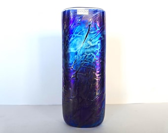 Könnte beinhalten: Eine hohe, zylindrische Glasvase mit strukturierter, irisierender Oberfläche. Die Vase wechselt von tiefem Lila zu leuchtendem Blau, mit einem Rissmuster. Auf der Oberseite der Vase befindet sich ein kleines Etikett mit der Aufschrift "GOZO".