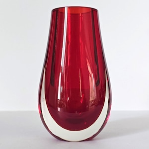 May include: A red glass vase in a teardrop shape with a white base. The vase has a glossy finish and a smooth surface. The red glass is translucent. A decorative item for the home, suitable for displaying flowers.