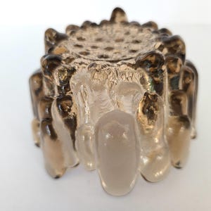 May include: A clear glass candle holder with a textured, frosted finish. The holder has a circular top with a raised, textured design and a base with a series of rounded, textured protrusions.