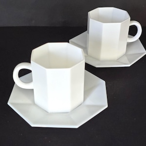 Peut inclure: Deux tasses à café blanches de forme géométrique avec des soucoupes assorties. Les tasses et les soucoupes ont un design angulaire moderne. Les tasses ont une anse et reposent sur une surface noire.