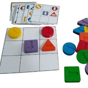 Op de afbeelding: Een kleurrijk educatief spel met een raster en verschillende vormen, waaronder cirkels, vierkanten en driehoeken, elk met een pijlsymbool. De set bevat kaarten met pijlenpatronen. De vormen zijn geel, rood, paars, oranje, groen en blauw.
