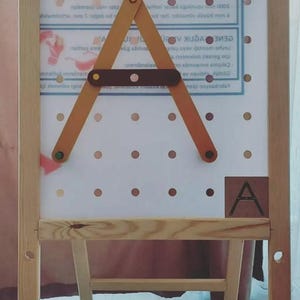 May include: Wooden easel with a white pegboard and a wooden A-shaped element. The easel has a small shelf and a wooden frame. The letter "A" is printed on a small wooden square.