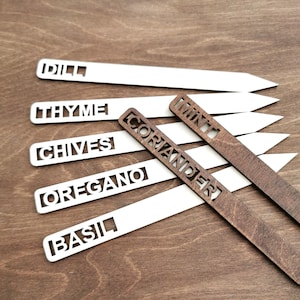Op de afbeelding: Houten kruidentuinmarkers met uitgesneden letters. De markers zijn wit en bruin, met namen als "Dille", "Tijm" en "Koriander". De markers zijn gerangschikt op een houten oppervlak, ideaal voor een kruidentuin.