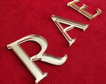 May include: Three gold-coloured letters, R, A, and E, are arranged in a row on a red velvet surface. The letters are made of a reflective material, possibly acrylic or metal.