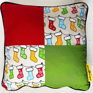 May include: A square Christmas cushion with a patchwork design. It features a red square, a green square, and two squares with a cream background and colourful Christmas stockings. The cushion has a black border.