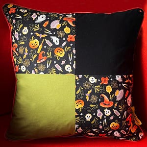 May include: Square Halloween cushion with a patchwork design. The cushion features a black and orange patterned fabric with pumpkins, bats, and witch hats, a solid black square, and a solid green square. The cushion has a red border.