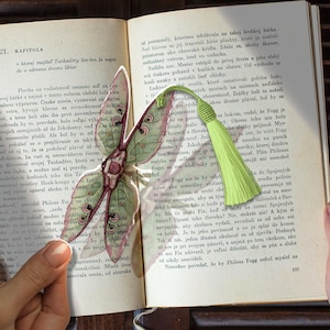 May include: A decorative bookmark featuring a detailed, die-cut butterfly with a green and purple color scheme. The butterfly is placed over an open book with text, and a green tassel hangs from the top.