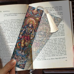 May include: A colorful bookmark featuring an illustration of a deity with a staff, adorned with intricate details. The bookmark has a cream-colored tassel and is placed on a book with text.