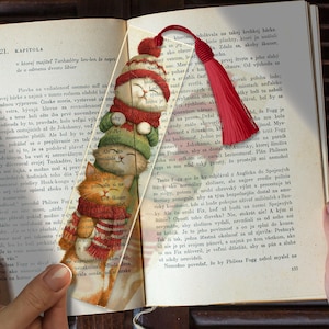 May include: A rectangular clear bookmark featuring a vertical stack of three illustrated cats wearing winter hats and scarves, topped with a red tassel. The bookmark is placed inside an open book with printed text visible through the transparent material.