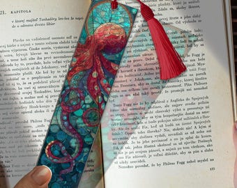 May include: A decorative bookmark featuring an octopus design in shades of red, teal, and blue. The bookmark has a red tassel and is placed on a book with text.
