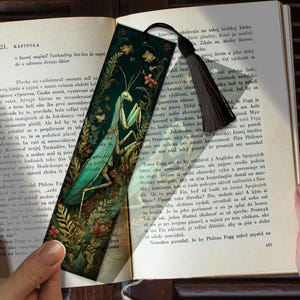 May include: A bookmark with a detailed illustration of a praying mantis in a dark green and gold floral setting. It has a black tassel and is set against an open book. The bookmark is approximately 18 cm long.