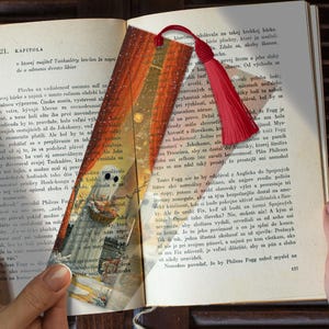 Może przedstawiać: Ozdobna zakładka do książki z czerwonymi frędzlami, spoczywająca na stronie książki. Zakładka przedstawia ilustrację w stylu vintage statku żeglującego po morzu, z ciepłą paletą barw.