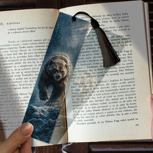 Pode incluir: Marcador de livros com uma ilustração de um urso num cenário florestal. Possui um borla preta e está colocado num livro aberto com texto.