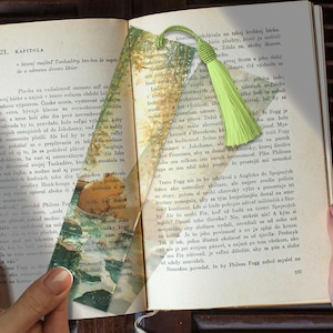 Peut inclure: Un signet transparent avec un pompon vert clair, placé dans un livre ouvert. Le signet présente un paysage avec des arbres et une rivière. Les pages du livre affichent du texte tchèque.