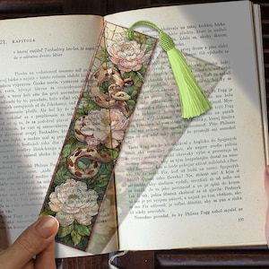 May include: A decorative bookmark featuring a detailed illustration of a snake coiled around white and pink flowers. The bookmark has a green tassel and is set against the backdrop of an open book.