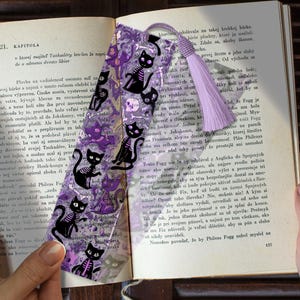 May include: A bookmark featuring a purple background with black cat skeletons, skulls, and bones. A lavender tassel hangs from the top. The bookmark is placed on an open book with text.