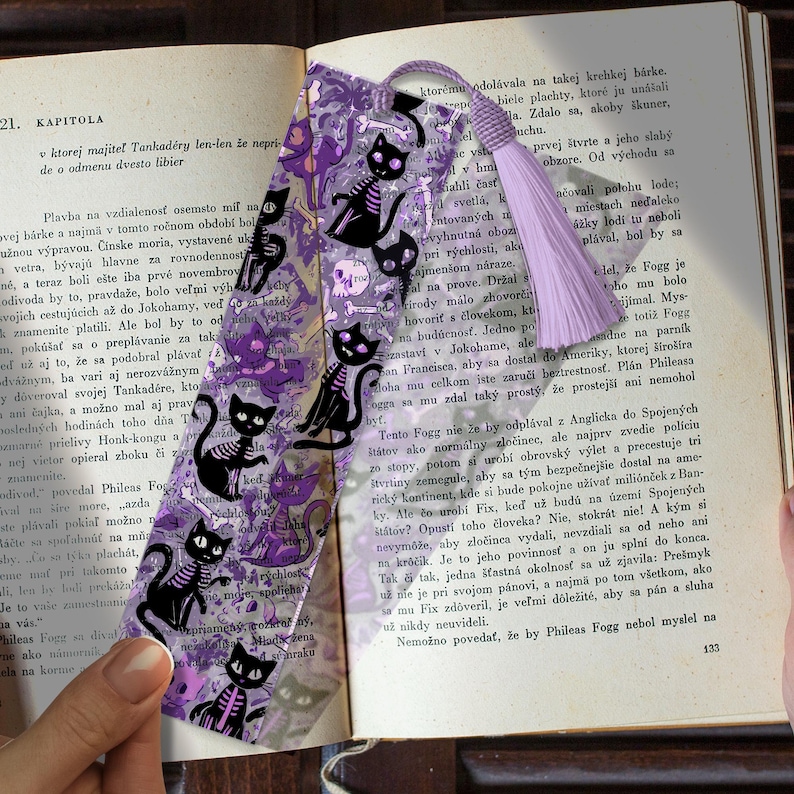 May include: A bookmark featuring a purple background with black cat skeletons, skulls, and bones. A lavender tassel hangs from the top. The bookmark is placed on an open book with text.