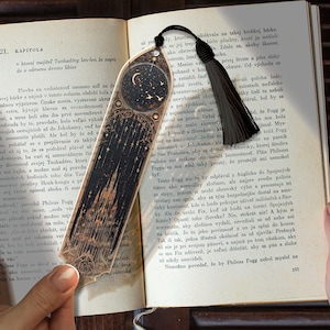 May include: A detailed bookmark with a black tassel. The design features a castle, stars, and a crescent moon, with a dark background and gold detailing. Ideal for marking pages in a book.