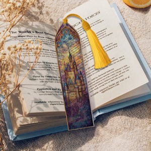 May include: A decorative bookmark with a castle illustration and a yellow tassel. The bookmark is in an open book. The castle design features blue, purple, and yellow hues. The book's pages are visible.
