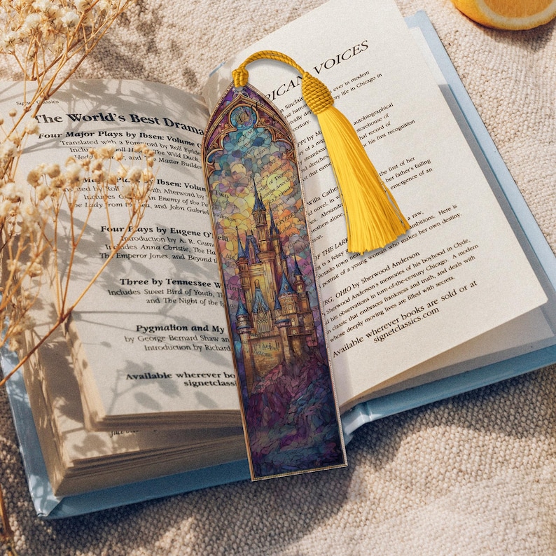 May include: A decorative bookmark with a castle illustration and a yellow tassel. The bookmark is in an open book. The castle design features blue, purple, and yellow hues. The book's pages are visible.