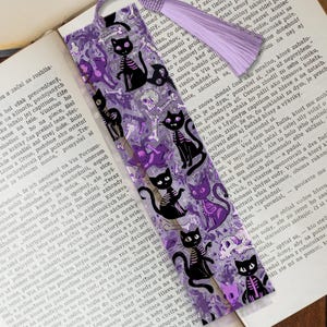 May include: A bookmark with a purple tassel features a repeating pattern of black cat skeletons, skulls, and bones on a translucent purple background. The bookmark is placed on an open book.