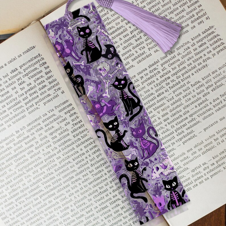 May include: A bookmark with a purple tassel features a repeating pattern of black cat skeletons, skulls, and bones on a translucent purple background. The bookmark is placed on an open book.