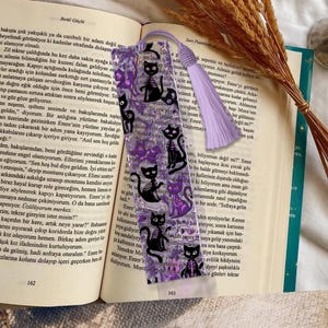 May include: A bookmark featuring a repeating pattern of black cat skeletons, purple accents, and text. The bookmark has a purple tassel and is placed in an open book.