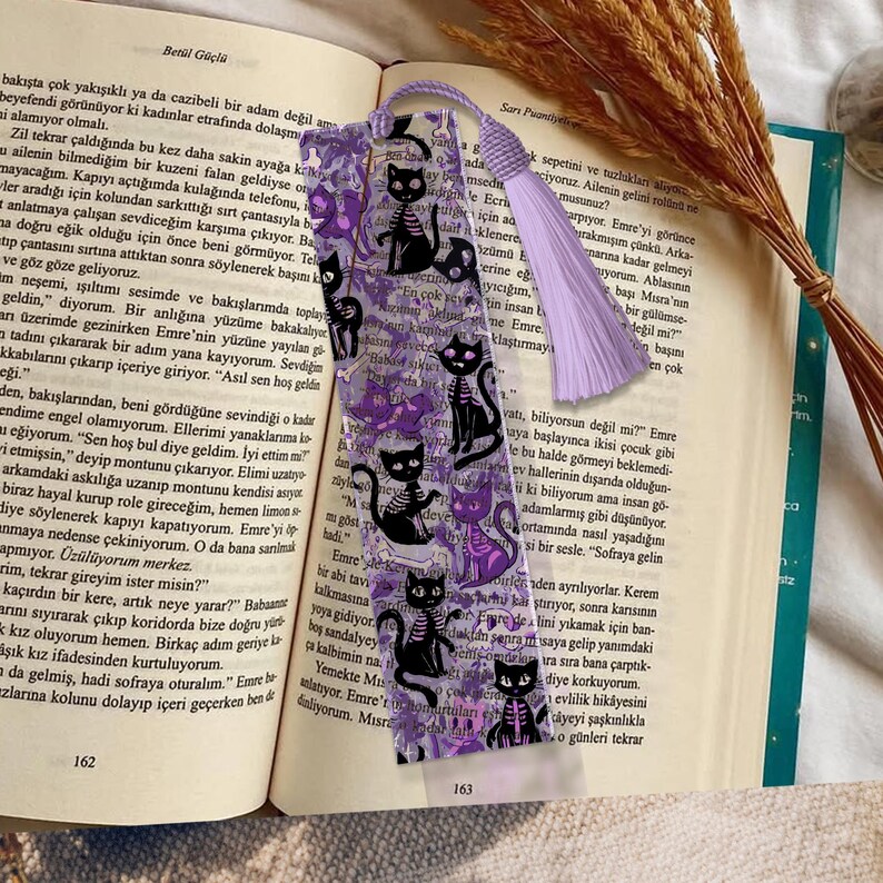 May include: A bookmark featuring a repeating pattern of black cat skeletons, purple accents, and text. The bookmark has a purple tassel and is placed in an open book.