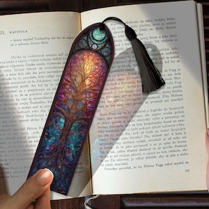Op de afbeelding: Een kleurrijke boekenlegger met een glas-in-lood ontwerp met een boom. De boekenlegger heeft een zwarte kwast en ligt op een open boek.