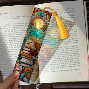 May include: A decorative bookmark with a yellow tassel, showing a hedgehog reading books, set against a stained glass design. The bookmark is inserted into a book, with the text "Will he marry Lodi?" visible.