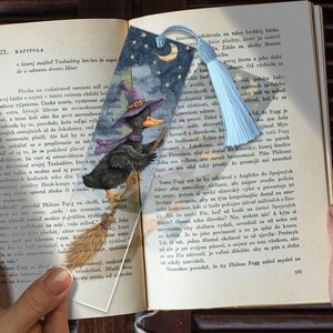 May include: A rectangular bookmark featuring a dark blue night sky with a crescent moon and stars. A black duck wearing a purple witch hat and scarf flies on a brown broomstick. The bookmark has a light blue tassel attached to the top hole.