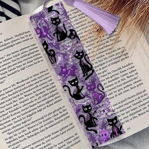 May include: A purple and black bookmark featuring a repeating pattern of black cat skeletons, purple cat silhouettes, and bones. The bookmark has a purple tassel and a purple ribbon.