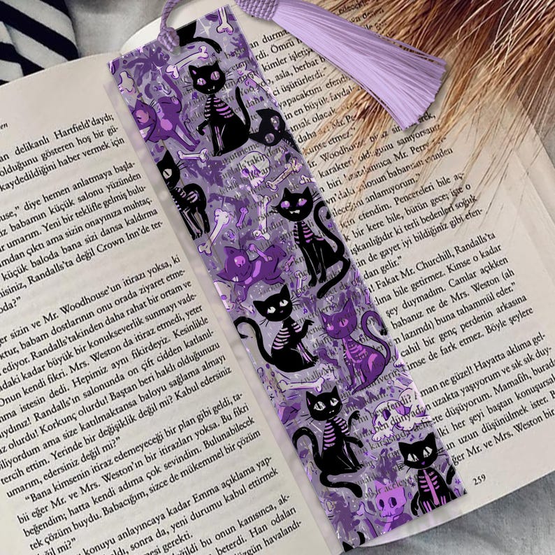 May include: A purple and black bookmark featuring a repeating pattern of black cat skeletons, purple cat silhouettes, and bones. The bookmark has a purple tassel and a purple ribbon.
