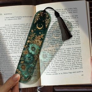 May include: A decorative bookmark with a dark teal background, featuring a crescent moon, stars, and floral accents. A black tassel hangs from the top. The bookmark is placed in an open book.