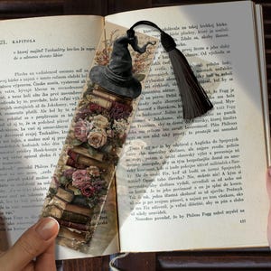 Op de afbeelding: Een rechthoekige boekenlegger met een stapel vintage boeken, bekroond met een donkergrijze heksenhoed en versierd met trossen roze en crèmekleurige rozen. Een zwarte kwast is bevestigd aan de bovenkant van de boekenlegger in een open boek.