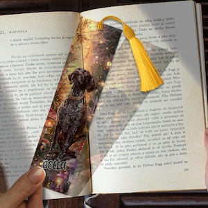 May include: Bookmark featuring a detailed illustration of a dog with a yellow tassel. The bookmark has the name "BELLA" and is set against a backdrop of a book page with text.