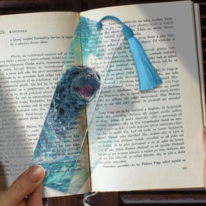 Pode incluir: Um marcador de livros com uma borla azul e uma ilustração de foca. O marcador está colocado num livro aberto com texto numa língua estrangeira.