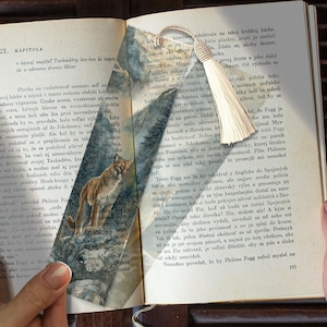 Pode incluir: Marcador de livros com uma ilustração de um puma, contra um fundo de paisagem montanhosa e texto de livro. Possui uma borla creme.