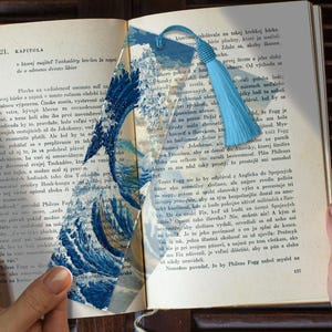 May include: A translucent bookmark featuring a blue and white wave design, with a matching blue tassel. The bookmark is inserted into a book with text visible, creating a decorative and functional accessory.