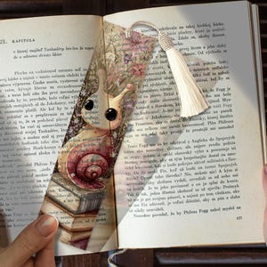 May include: A rectangular paper bookmark featuring a whimsical illustration of a snail with large black eyes, resting on a stack of books amidst a floral background. The bookmark is topped with a cream-coloured decorative tassel attached by a cord.