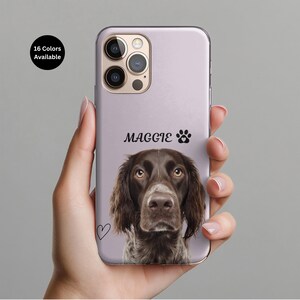 May include: A phone case featuring a dog portrait with the name "Maggie" and a paw print. The case is light purple with a heart graphic and the text "16 Colors Available". The dog has brown fur and eyes.