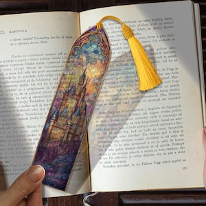 Peut inclure: Marque-page décoratif avec un château de style vitrail aux teintes bleues, violettes et orangées. Il est orné d'un pompon jaune et se trouve dans un livre ouvert.
