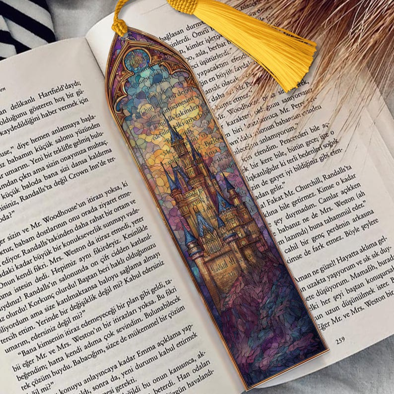 May include: A decorative bookmark featuring a castle design in a stained-glass style, with a yellow tassel. The bookmark is positioned on a book, adding to its aesthetic.