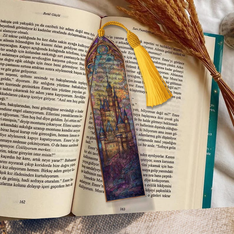 May include: A decorative bookmark featuring a castle illustration, a yellow tassel, and a patterned top. The bookmark is in an open book with text on the pages.