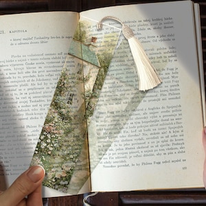 Op de afbeelding: Een rechthoekige papieren boekenlegger met een gedrukte illustratie van een huisje omringd door weelderig groen gebladerte en bloemen. De boekenlegger heeft een crèmekleurig koordkwastje aan de bovenkant en ligt in een open boek.