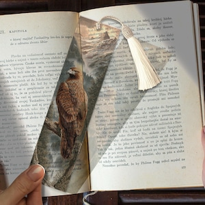 Pode incluir: Marcador de livros decorativo com uma ilustração detalhada de um falcão marrom empoleirado em um galho, em um fundo de floresta e montanhas. O marcador de livros tem uma borla creme.