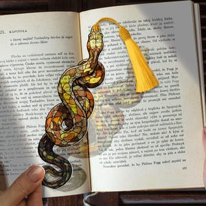 Può includere: Un segnalibro a forma di serpente, con un design a vetrata colorata nei toni del marrone, arancione e giallo. Ha un nappa gialla ed è appoggiato su un libro aperto.