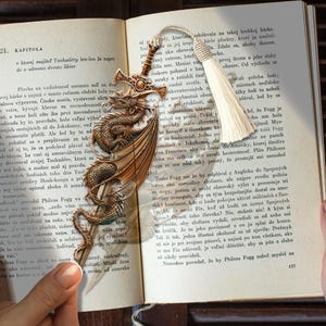 May include: Ornate metal bookmark featuring a dragon wrapped around a sword. The bookmark has a tassel and is set on the pages of a book with text.