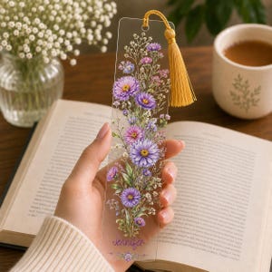 May include: A clear rectangular bookmark featuring a printed illustration of purple and pink wildflowers with green stems and leaves. The name Jennifer is printed in purple script at the bottom. A gold-coloured tassel is attached to the top hole.